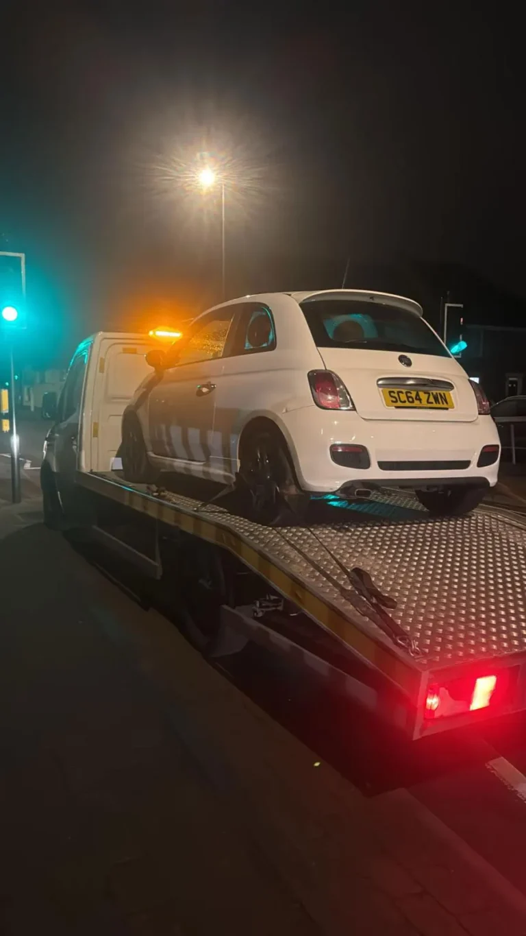 24/7 breakdown recovery flatbed tow truck transporting a white Fiat 500 during a nighttime roadside vehicle recovery.