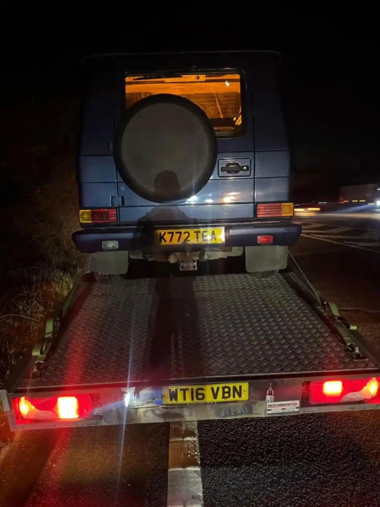 Nighttime rear view of a dark blue Mercedes G-Class SUV with spare tire mounted on the tailgate, loaded onto a flatbed recovery truck on a roadside at night.