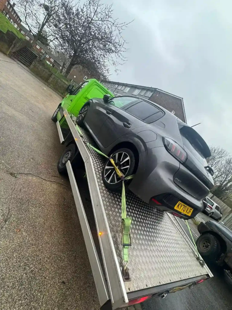 A grey Peugeot 208 secured with straps on a green flatbed recovery truck, parked on a residential street on an overcast day.