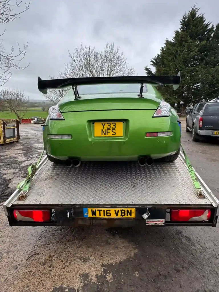 Rear view of a green Nissan 370Z with a large rear spoiler and quad exhaust, loaded onto a flatbed recovery truck on an overcast day.