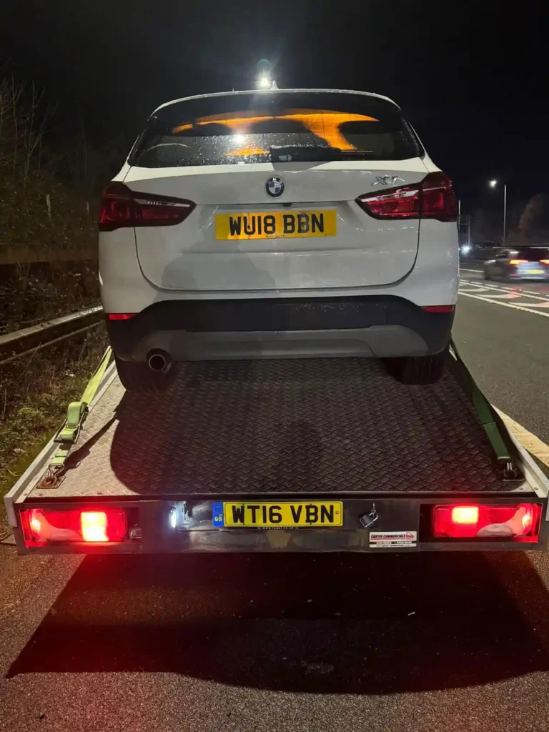 Nighttime rear view of a white BMW X1 loaded onto a flatbed recovery truck parked on the roadside.