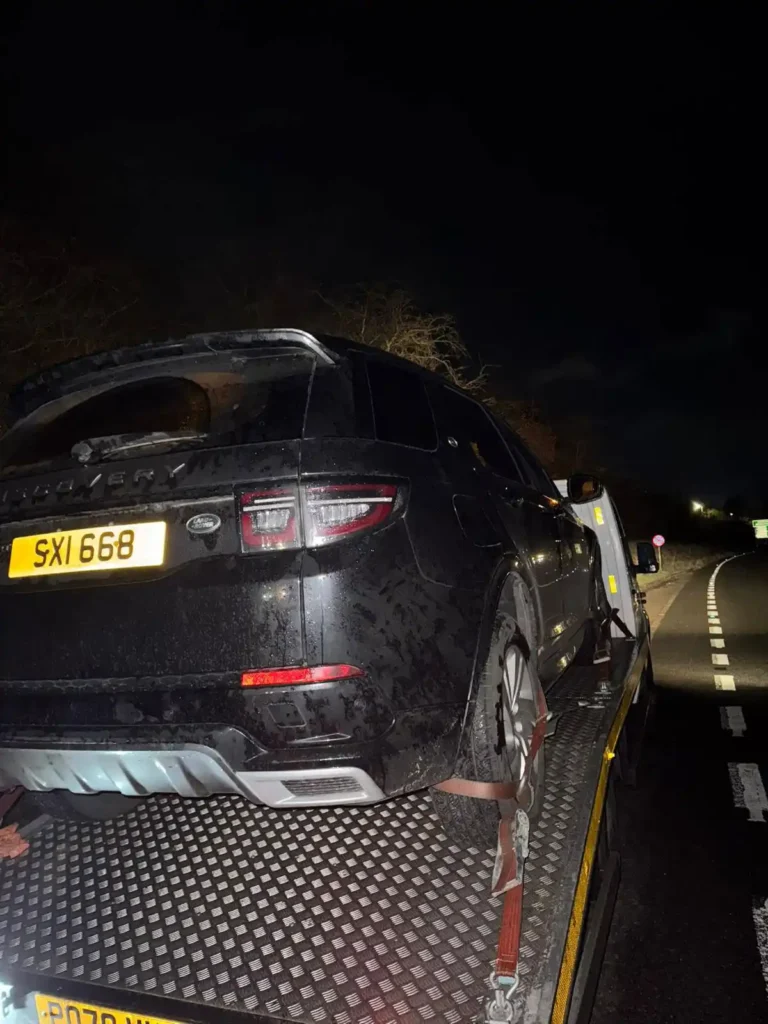 Nighttime rear view of a wet black Land Rover Discovery strapped onto a flatbed recovery truck on a country road.