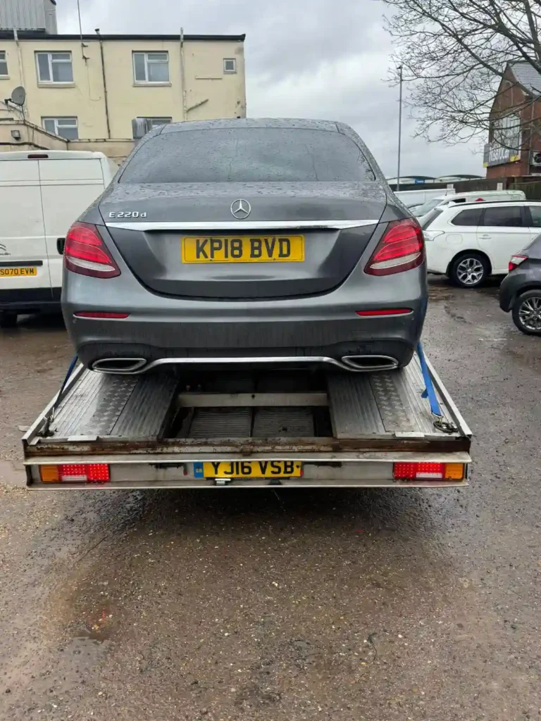 Rear view of a grey Mercedes E220d strapped onto a flatbed recovery truck in a parking area on an overcast day.